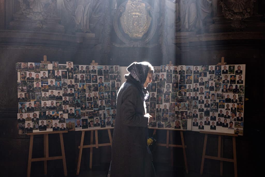 A woman prays as she walks past a bulletin board showing portraits of those killed by invading Russian forces in Lviv, Ukraine, on 3.18.22. Ukraine mourns every loss, while recognizing that there are more to come. Two years later, it’s impossible to imagine what this board would look like if it reflected an updated death toll. © Alex Kent